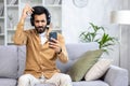 Happy young Indian man sitting at home on sofa wearing headphones and using mobile phone, relaxing, listening to music Royalty Free Stock Photo