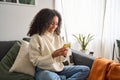 Happy young latin woman sitting on sofa using phone in living room. Royalty Free Stock Photo