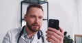 Happy young male doctor consulting remote patient on self isolation using smart phone video call chat app at office. Royalty Free Stock Photo