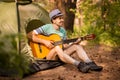 Happy young man camping and strum a guitar instrumental music to relax against background of forest sunset.