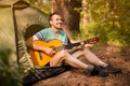 Happy young man camping and strum a guitar instrumental music to relax against background of forest sunset.