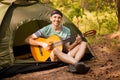 Happy young man camping and strum a guitar instrumental music to relax against background of forest sunset Royalty Free Stock Photo