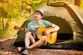 Happy young man camping and strum a guitar instrumental music to relax against background of forest sunset. Wilderness Travel
