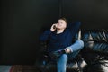 Happy young man in casual clothes lying on a black leather sofa and talking on the phone against a dark wall, sideways with a Royalty Free Stock Photo