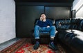 Happy young man in casual clothes sitting on a black leather couch, talking and on the phone and smiling looking into the camera. Royalty Free Stock Photo