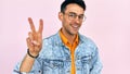 Happy young man in casual stylish clothes, transparent eyeglasses showing victory gesture, isolated on pink studio background. Royalty Free Stock Photo