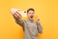 Happy young man in glasses and shirt is taking a selfie on a yellow background, looking into the camera of a smartphone, smiling Royalty Free Stock Photo