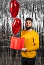 Happy young man with heart-shaped balloon and red gift box on foil background. Valentine's Day celebration Royalty Free Stock Photo