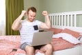 Happy young man in his home bedroom sitting with a laptop on his legs and hands up high