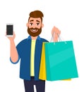 Happy young man holding shopping bags. Male character carrying colourful bags. Person showing cell, mobile or smartphone in hand.