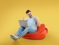 Happy young man with laptop on bean bag chair, yellow background Royalty Free Stock Photo