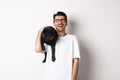 Happy young man laughing carefree, holding cute black dog, pug breed, on shoulder and having fun, standing over white