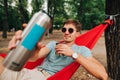 Happy young man lies in a red hammock tied to a tree in the woods with a cup of drink in his hands, looks into the camera with a Royalty Free Stock Photo