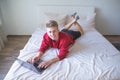 Happy young man lying on bed in a cozy bright room using a laptop, looking into the camera