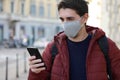 Happy young man with protective mask looks his smartphone in street. Student guy with face mask using mobile phone outdoors Royalty Free Stock Photo