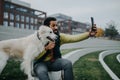 Happy young man resting and taking selfie with his dog outdoors in city park, during cold autumn day. Royalty Free Stock Photo