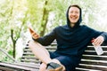 Happy Young man runner in hoodie sitting on bench and relaxing after sport training, using mobile phone. Holding water Royalty Free Stock Photo