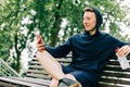 Happy Young man runner in hoodie sitting on bench and relaxing after sport training, using mobile phone. Holding water Royalty Free Stock Photo