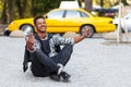 Happy young man seated down on pavement road with crossed legs,  on a yellow blurred taxi background. Royalty Free Stock Photo