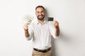 Happy young man showing his credit card and money dollars, smiling satisfied, standing over white background Royalty Free Stock Photo