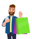 Happy young man showing shopping bags. Person holding a mobile, cell, smart phone in hand. Modern lifestyle, digital technology.