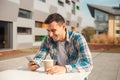 Happy young man sitting outdoors looking in his phone and holding cup of coffee