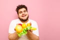 Happy young man standing on pink background with fruits and lettuce in hands, looking in camera and smiling, wearing a white t- Royalty Free Stock Photo