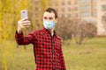 Happy young man in sterile face mask making self photo or video call with mobile phone show hello gesture walking by street Royalty Free Stock Photo