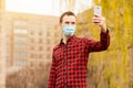 Happy young man in sterile face mask making self photo or video call with mobile phone show hello gesture walking by street Royalty Free Stock Photo