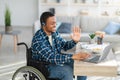 Happy young man in wheelchair communicating online, using laptop computer, waving at webcam at home Royalty Free Stock Photo