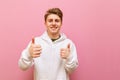 Happy young man in white casual clothes, looks into the camera with a smile on his face and shows thumbs up, isolated on pink Royalty Free Stock Photo