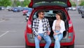 Happy young man and woman sitting on trunk of car outdoors while talking. Transportation, leisure, road trip, travel and Royalty Free Stock Photo