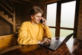 Happy young man working on laptop in the country house in a room with wooden interior and talking on the phone with a smile on his Royalty Free Stock Photo