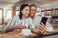 Happy young mixed race couple sitting at table having coffee while looking at something on smartphone in cafe. Loving Royalty Free Stock Photo