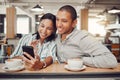 Happy young mixed race couple sitting at table having coffee while looking at something on smartphone in cafe. Loving Royalty Free Stock Photo