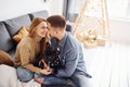Happy young modern couple sitting on the sofa at home with christmas tree with their cute dog Royalty Free Stock Photo