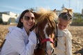 Happy young mother and daughter smiling looking at camera, hugging and posing with a little pony horse.