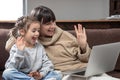 Happy young mother with daughter waving hands, looking at webcam using laptop for video call