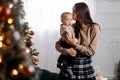 A happy young mother holds her little son in her arms against the backdrop of a beautifully decorated New Year tree and kisses him