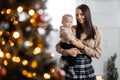 A happy young mother holds her little son in her arms against the backdrop of a beautifully decorated New Year tree and kisses him