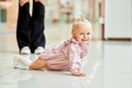 Happy young mother watch her cute baby crawling on floor at public place, shopping mall. Mom and her little daughter in Royalty Free Stock Photo