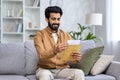 Happy young muslim man sitting on sofa at home, opening and reading received letter Royalty Free Stock Photo