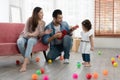 Happy young parents Caucasian father and Asian mother playing ukulele and singing with little kid girl in living room at new house