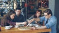 Happy young people are cutting large pizza with knives, talking and laughing while dining in cozy cafe. Modern lifestyle Royalty Free Stock Photo