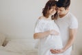 Happy young pregnant couple holding belly bump on white bed. Stylish pregnant family, mom and dad in white, relaxing at home and Royalty Free Stock Photo