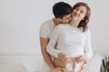 Happy young pregnant couple holding white little socks on belly bump on white bed. Stylish pregnant family, mom and dad in white, Royalty Free Stock Photo