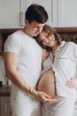 Happy young pregnant couple in white pajamas holding belly bump in kitchen in the morning. Stylish pregnant family, mom and dad Royalty Free Stock Photo