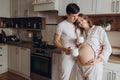 Happy young pregnant couple in white pajamas holding belly bump in kitchen in the morning. Stylish pregnant family, mom and dad Royalty Free Stock Photo