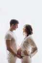 Happy young pregnant couple in white pajamas holding belly bump in morning light. Stylish pregnant family, mom and dad, relaxing Royalty Free Stock Photo