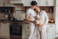 Happy young pregnant couple in white pajamas holding cups and talking in kitchen in the morning. Stylish pregnant family, mom and